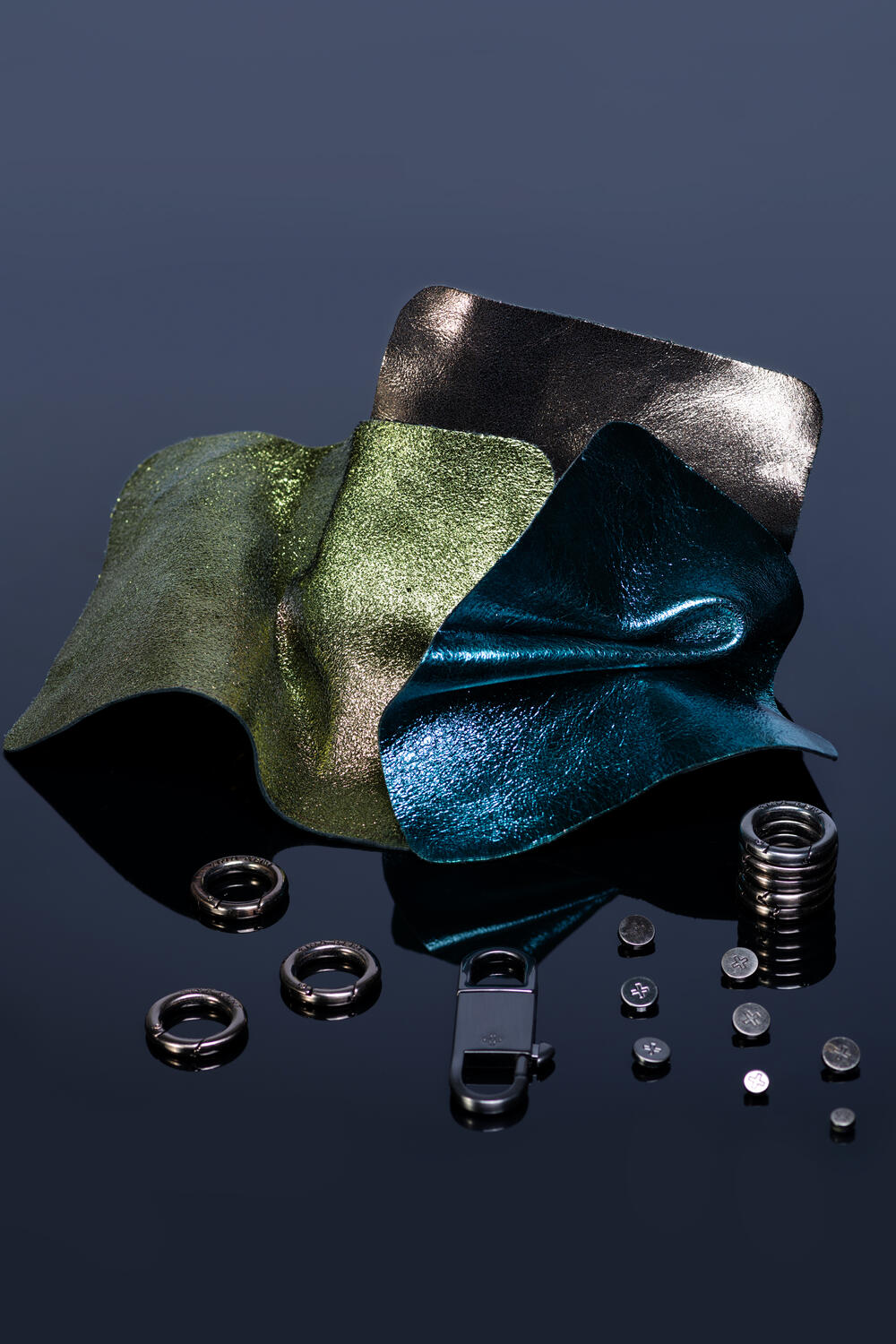 Samples of INA KENT metallic fabrics in green and blue are displayed with metal rings, a carabiner, and small metal discs on a reflective surface.