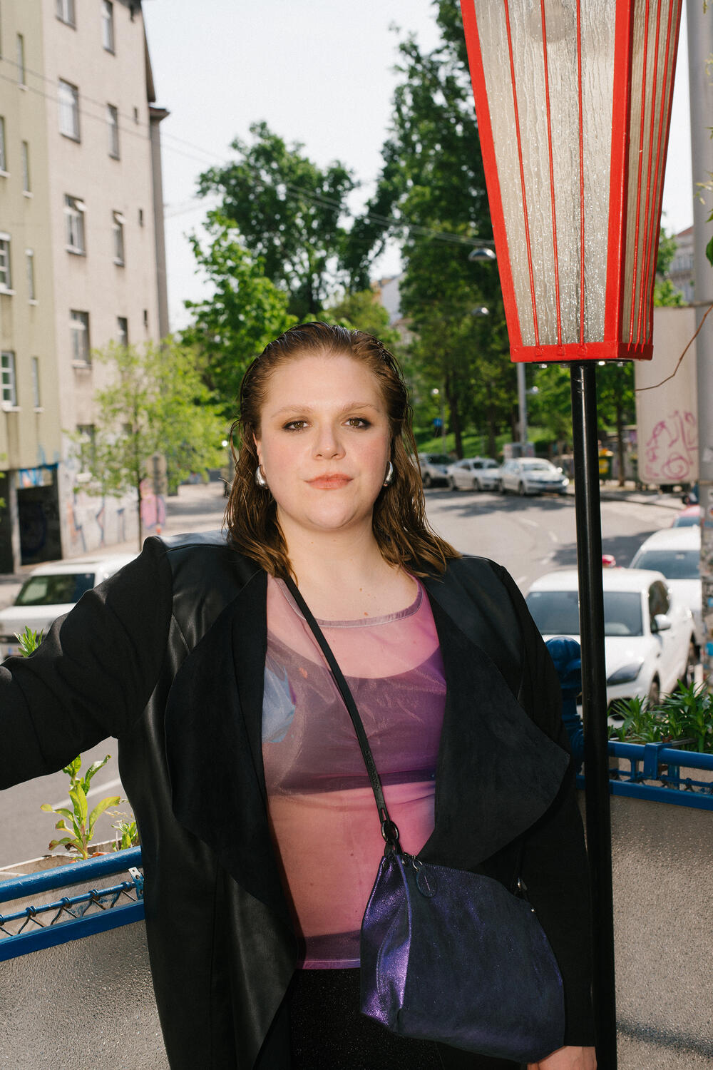 Eine Person steht im Freien. Sie trägt eine schwarze Jacke und ein lila Oberteil, ihr kurzes nasses Haar glänzt. Sie hält eine blaue Tasche von INA KENT in der Hand, während Stadtgebäude und Bäume eine ruhige Kulisse bilden.