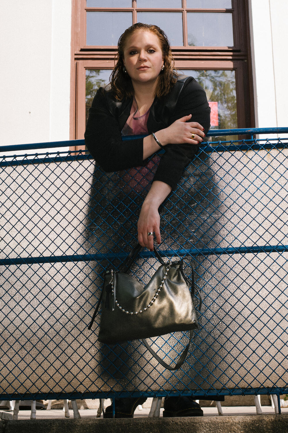 Eine Person lehnt über einem blauen Metallzaun und hält eine schwarze Handtasche von INA KENT in der Hand. Im Hintergrund ist ein Fenster zu sehen.