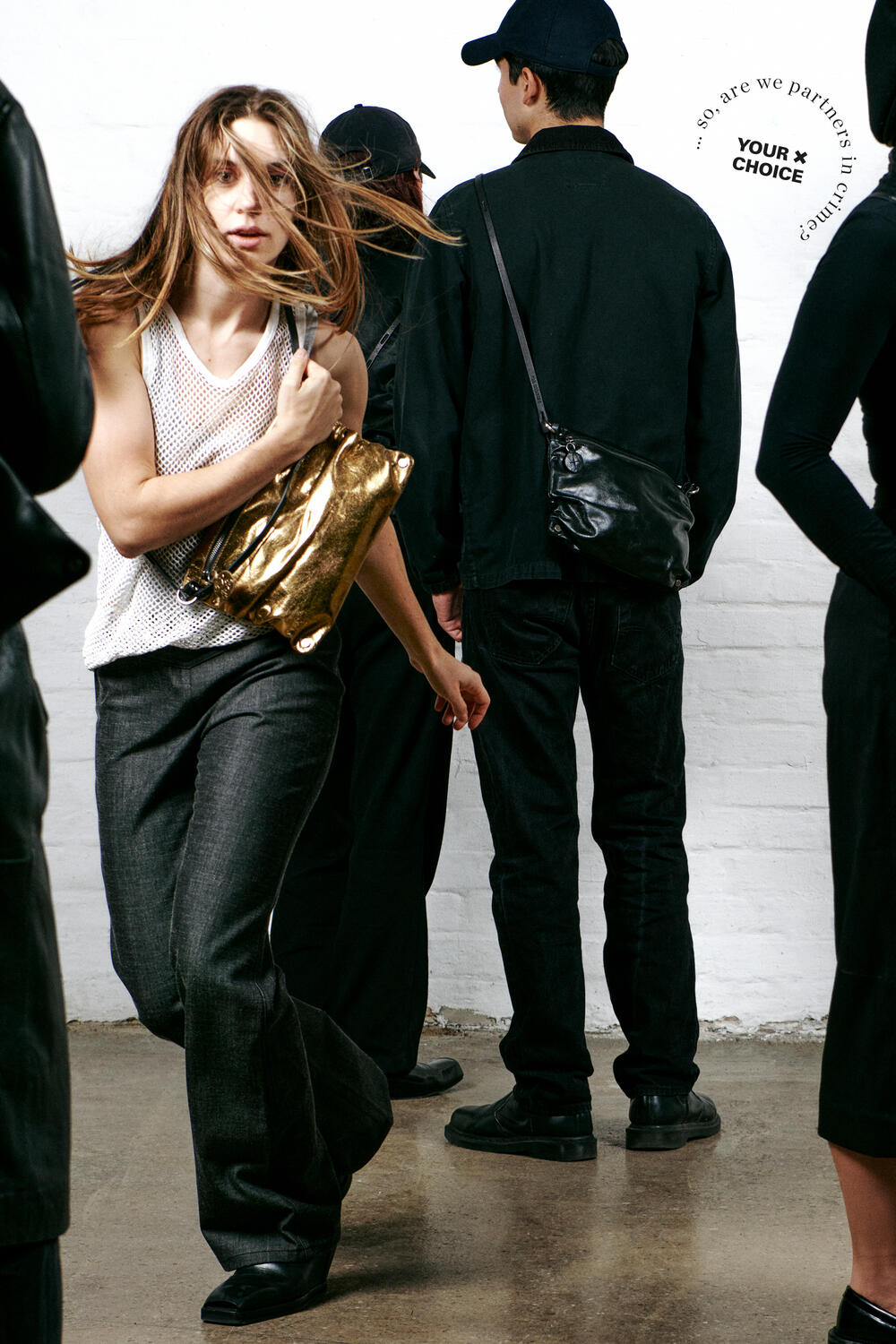 A person with long hair and a white mesh top walks energetically, holding a chic gold bag by INA KENT. Others in dark clothing stand in the background against a white brick wall.