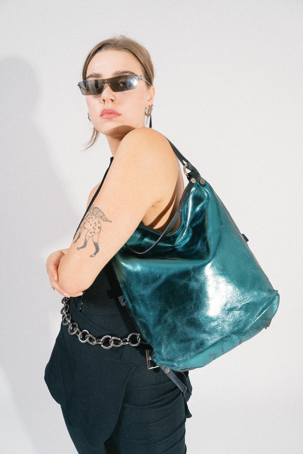 Person wearing sunglasses and black clothing with a tattoo on their arm, carrying an INA KENT metallic teal bag over their shoulder.