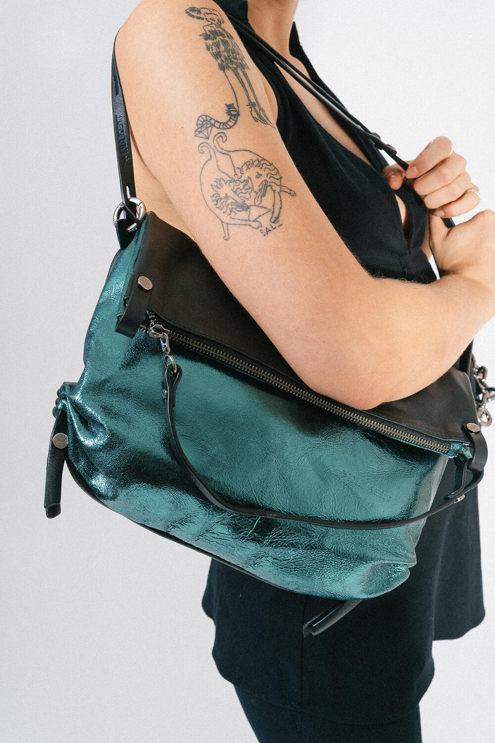 Person in a sleeveless top holding an INA KENT metallic green handbag with a zipper. Tattoo visible on arm.