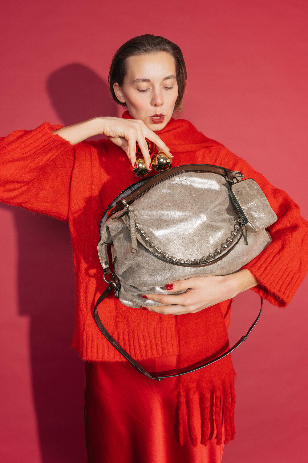 Frau in einem roten Outfit mit Pullover, Schal und Rock hält die INA KENT-Tasche DELA EDEN ed.1 in Crackled Anthra Leder vor ihrem Oberkörper. Sie schmückt die Tasche mit goldenen Weihnachtskugeln vor einem roten Hintergrund.