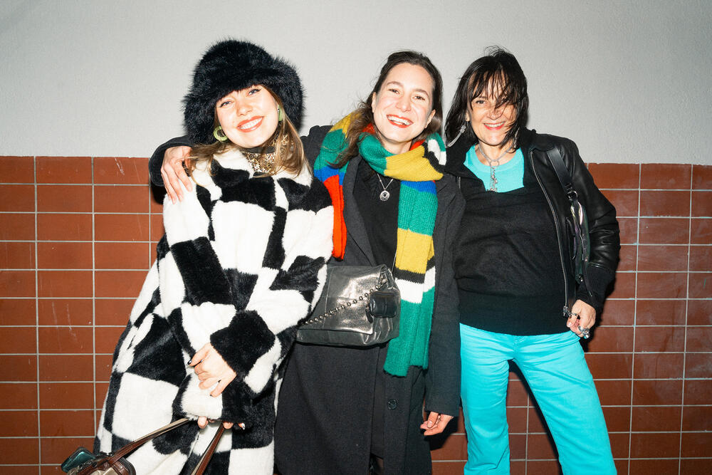 Three female-presenting people are standing next to each other, smiling. The person on the left is wearing a black and white patterned coat. The person in the middle is wearing a black coat, a colorful striped scarf and the bag “EILEEN ed.1” in crackled graphite. The person on the right is wearing a black leather jacket, a turquoise top and is holding the “DINKUM ed.2” bag in a shimmering design. 