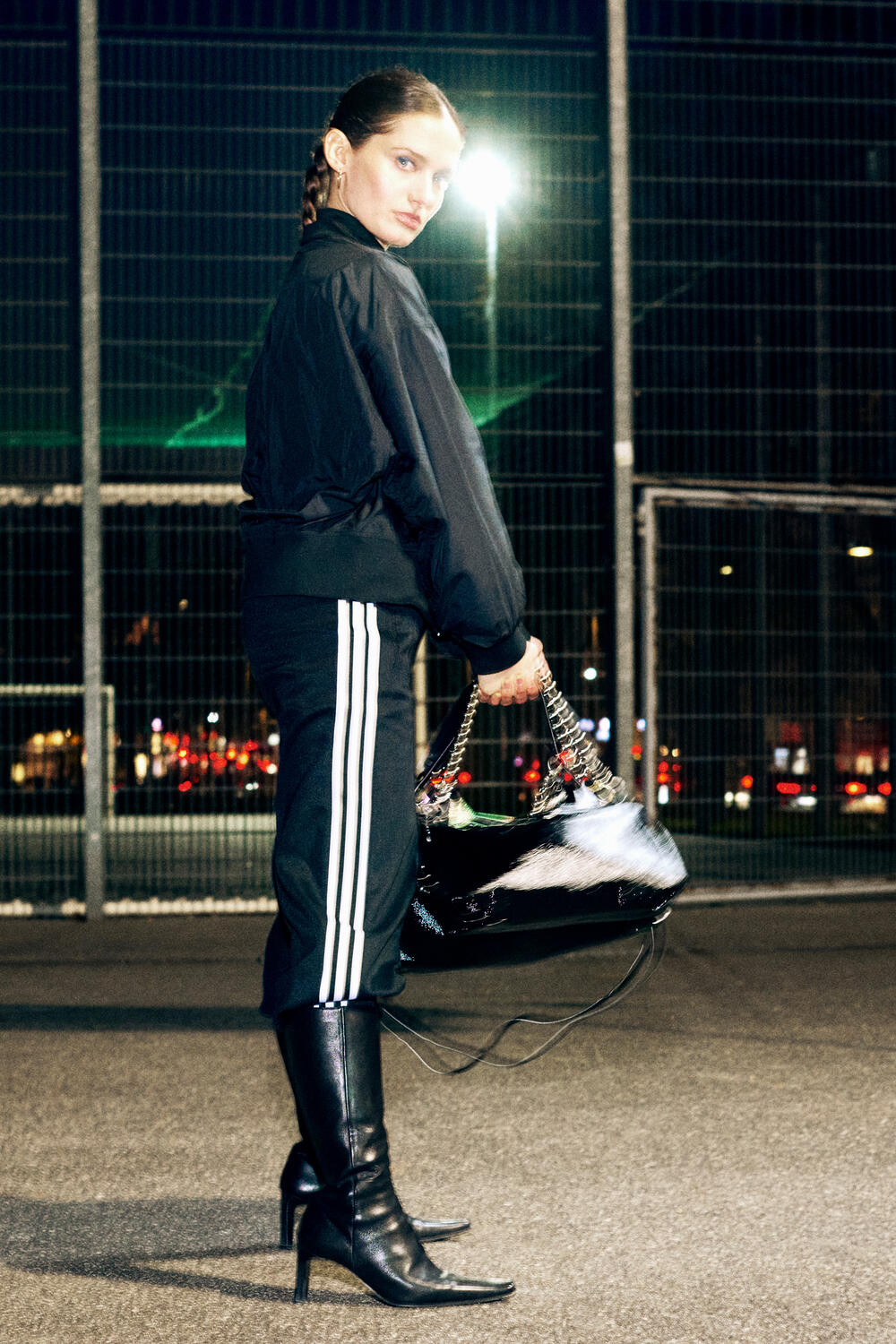 Eine Person in einer schwarzen Jacke, einer Trainingshose mit weißen Streifen und Stiefeln mit Absatz steht nachts im Freien und hält eine elegante schwarze Handtasche von INA KENT in der Hand.