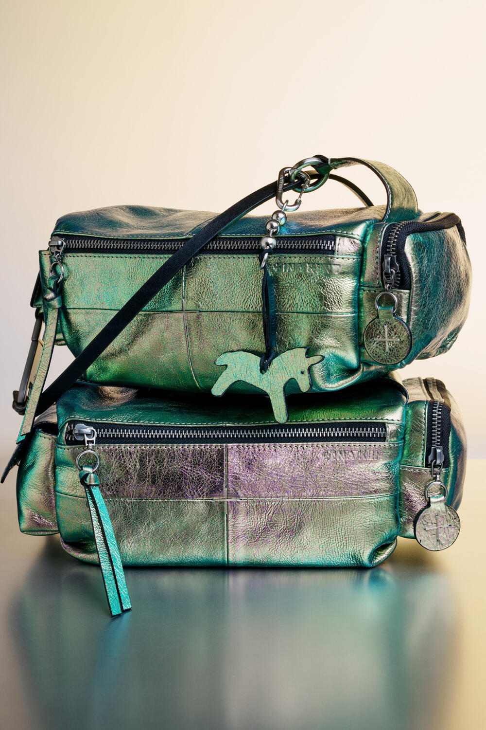Two metallic green leather INA KENT handbags with zippers and keychain charms, one stacked atop the other against a plain background.