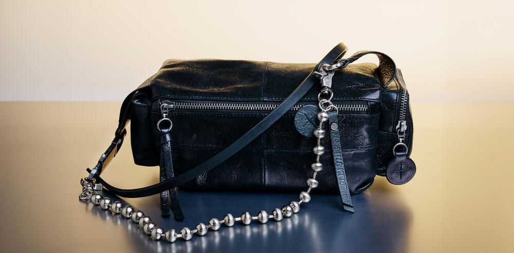 Eine schwarze Lederhandtasche von INA KENT mit Reißverschluss, Perlenkettenriemen und dekorativen runden Anhängern liegt elegant auf einer reflektierenden Oberfläche.