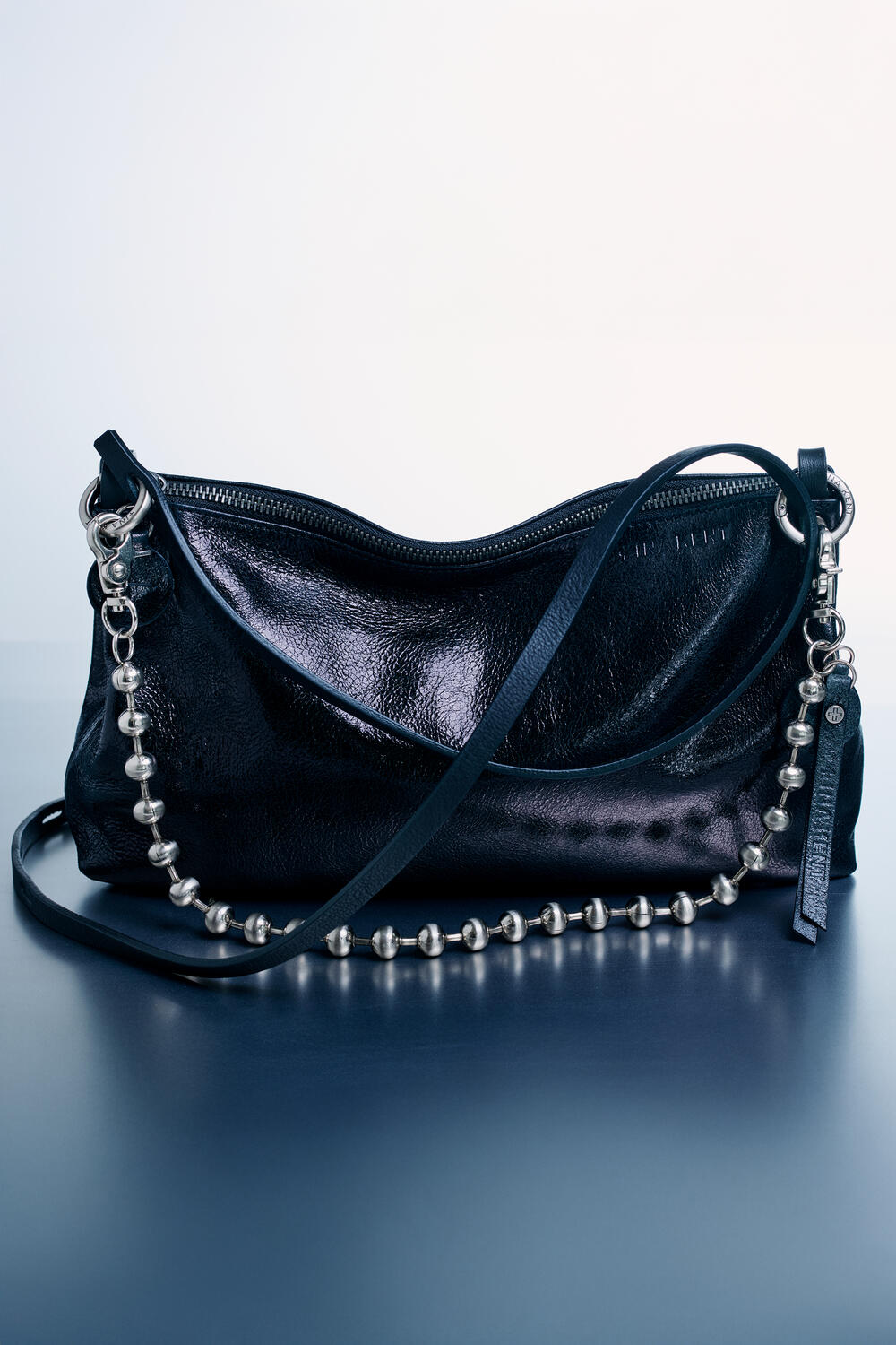 A black leather handbag by INA KENT with a shiny finish, featuring a zipper closure, a leather strap, and a decorative silver ball chain.