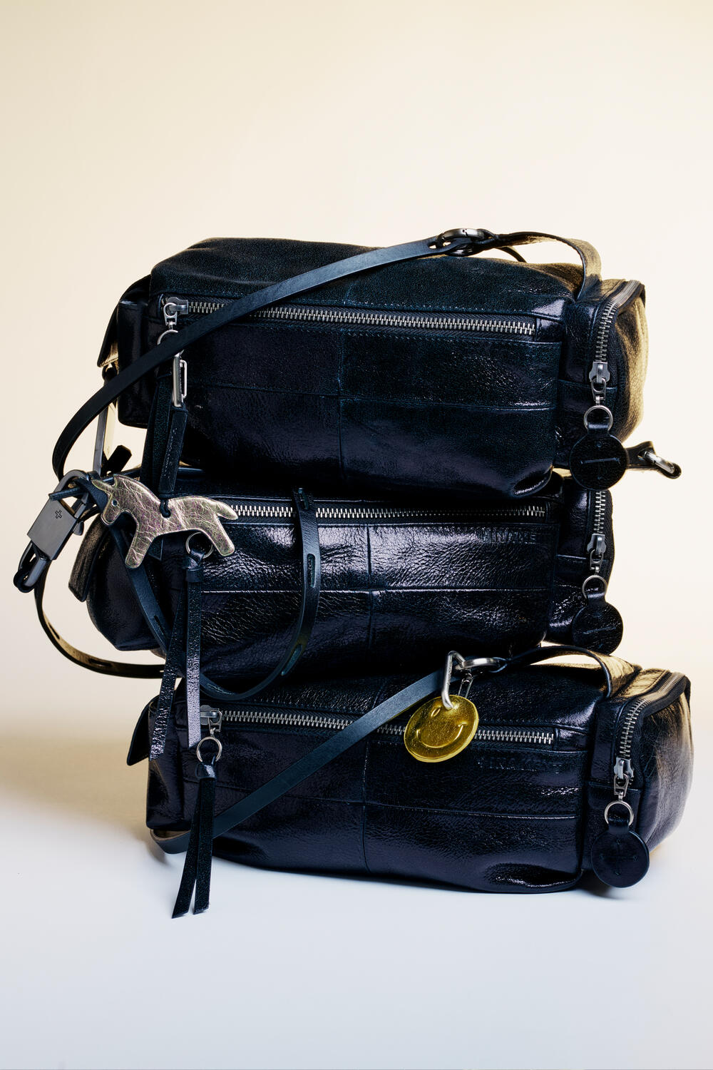 Drei schwarze Lederhandtaschen von INA KENT sind übereinander gestapelt, jede mit Reißverschlüssen und Schlüsselanhängern aus Metall in Form eines Pferdes und einer Münze.