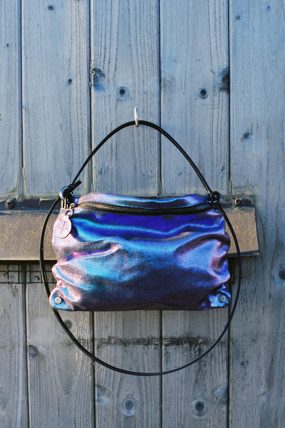 Eine metallisch schillernde Handtasche von INA KENT mit einem runden schwarzen Griff hängt an einer verwitterten Holztür.