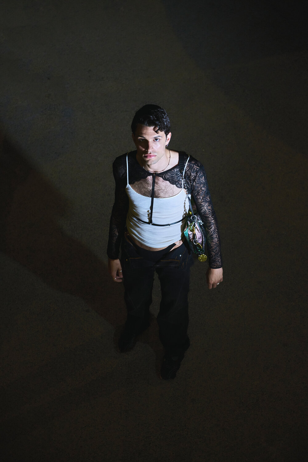 A person stands under a spotlight at night, wearing a white tank top over a black lace shirt, black pants, and holding an INA KENT patterned handbag, looking up at the camera.