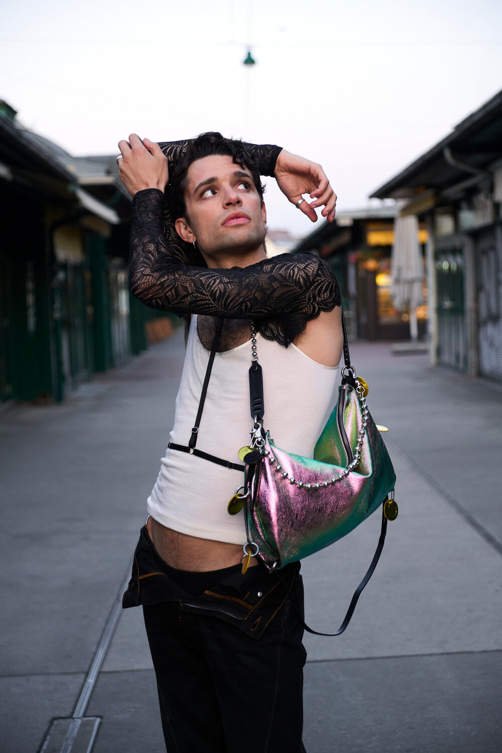 A person wearing a white tank top, black lace sleeves, and black pants poses outdoors with a shiny metallic INA KENT shoulder bag.