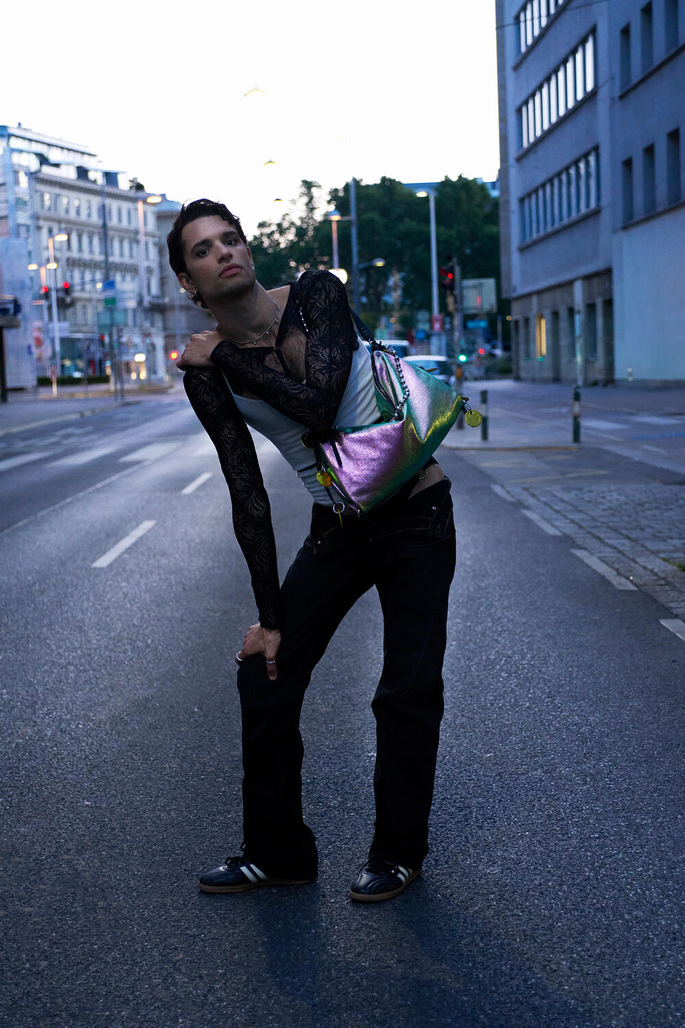 A person in black lace sleeves, black pants, and black sneakers poses on an empty city street at dusk, carrying a shiny iridescent INA KENT bag over their shoulder.