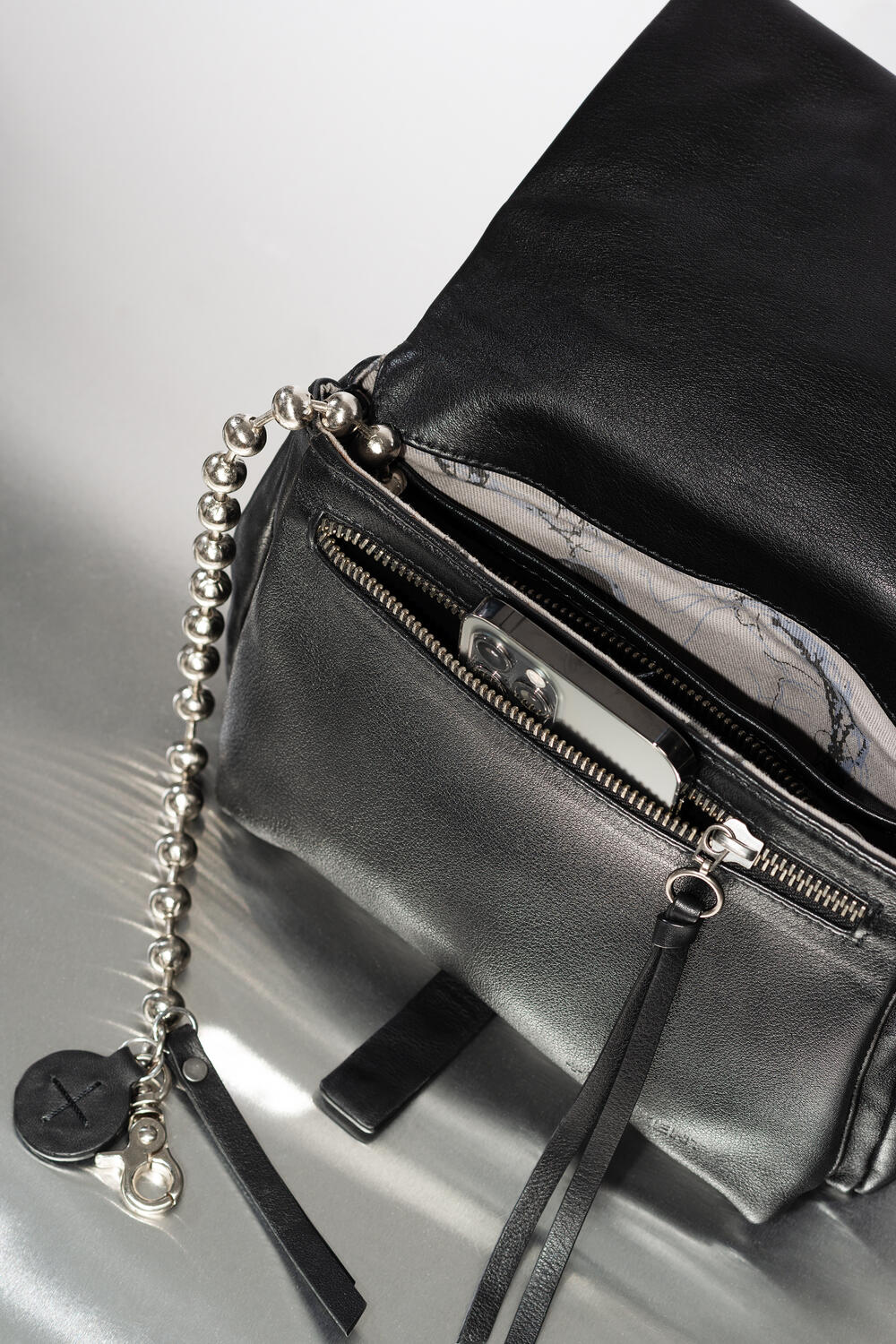 INA KENT Tasche aus schwarzem Leder mit metallischer Kugelkette, Reißverschluss und einer offenen Tasche, in der ein silbernes Smartphone steckt.
