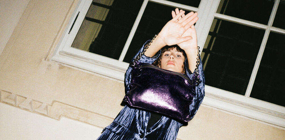 Person in a blue outfit holds up their arms, partially covering their face, with a shiny purple INA KENT bag slung over their shoulder, standing in front of a large window.
