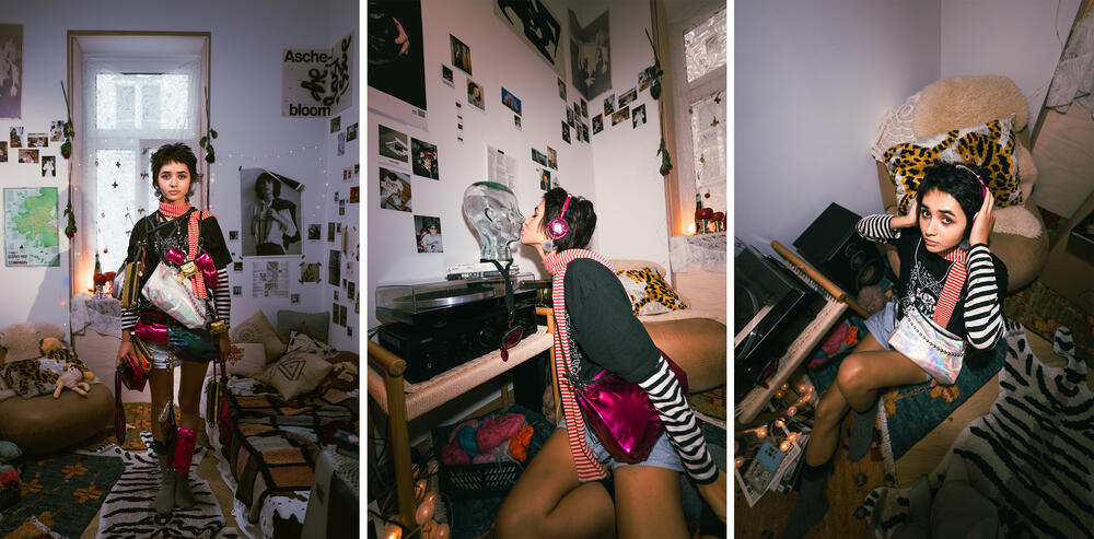 Eine junge Person mit kurzen Haaren und farbenfroher Kleidung posiert und hört Musik in einem dekorierten Schlafzimmer, das mit Postern, Fotos, verschiedenen persönlichen Gegenständen und einer INA KENT-Tasche gefüllt ist, die der lebhaften Szene eine stilvolle Note verleiht.