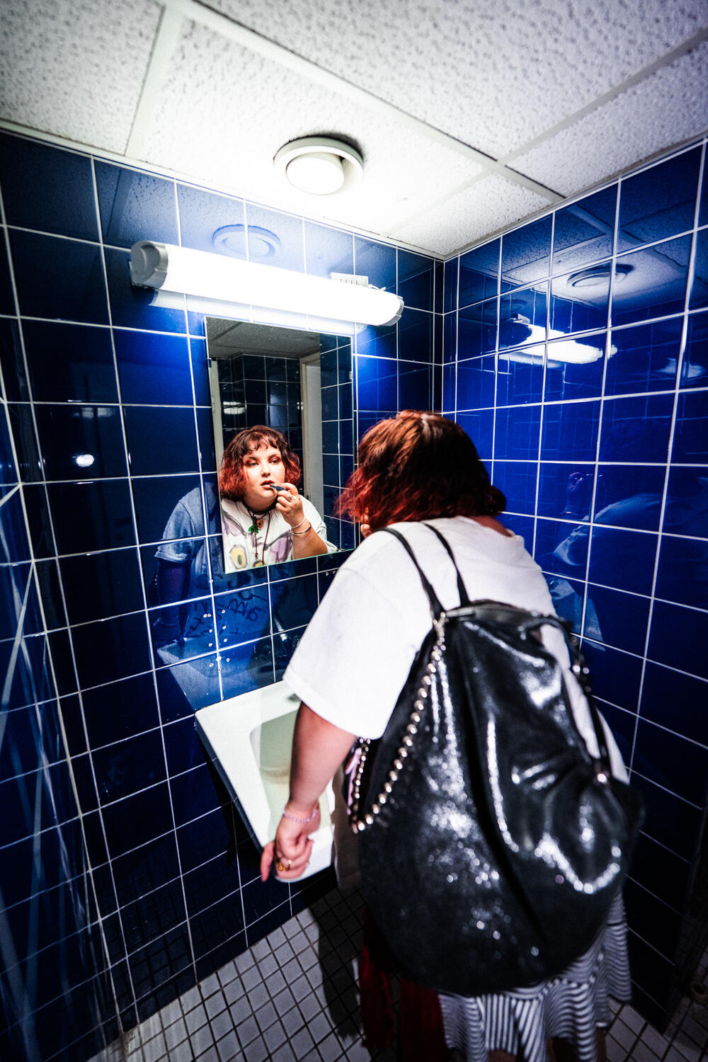 Person standing at a sink in a blue-tiled bathroom, looking into a mirror and applying makeup to their face, with an INA KENT bag resting nearby.