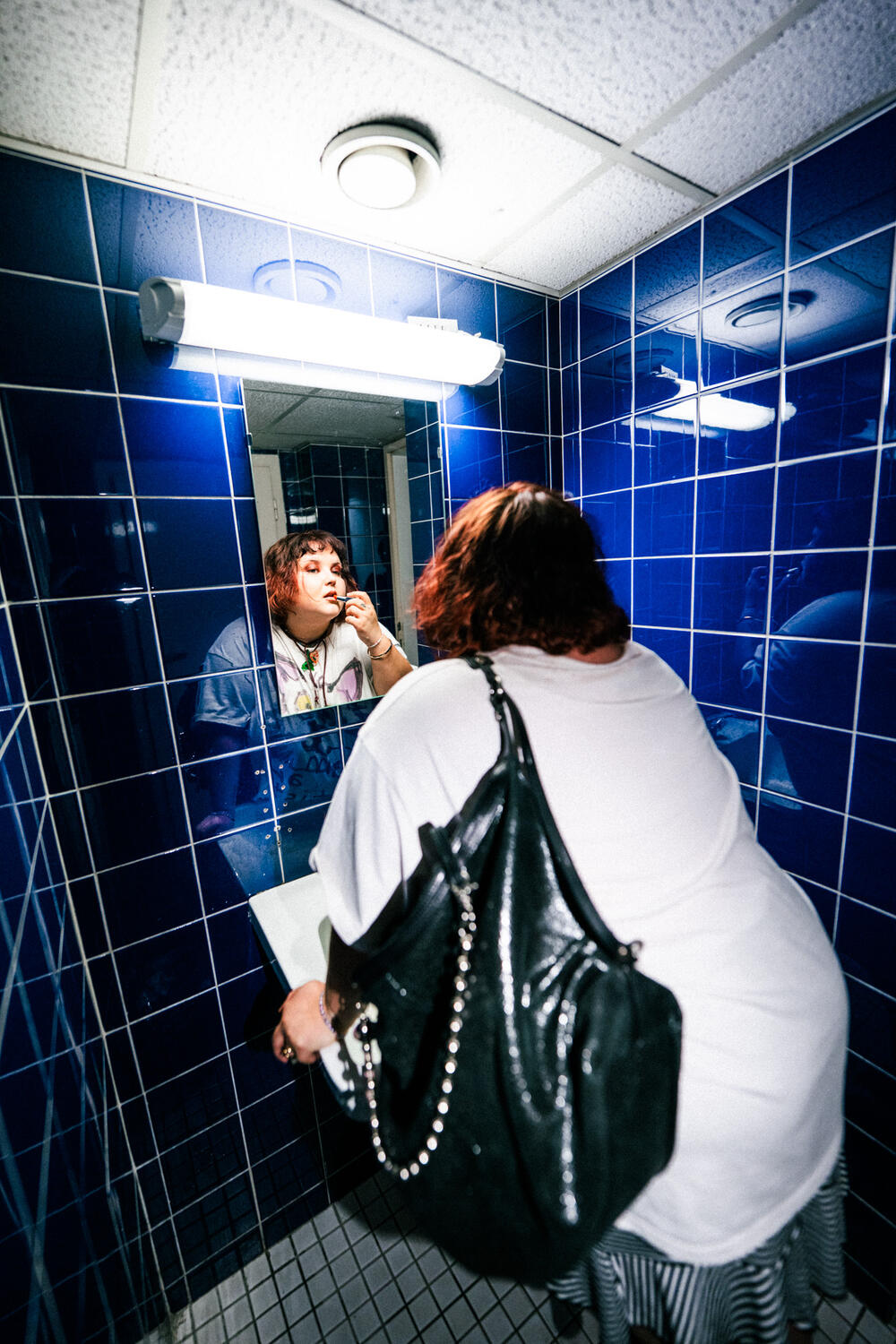 Person with an INA KENT black shoulder bag stands at a sink in a blue-tiled restroom, using a cotton swab on their face while looking in the mirror.
