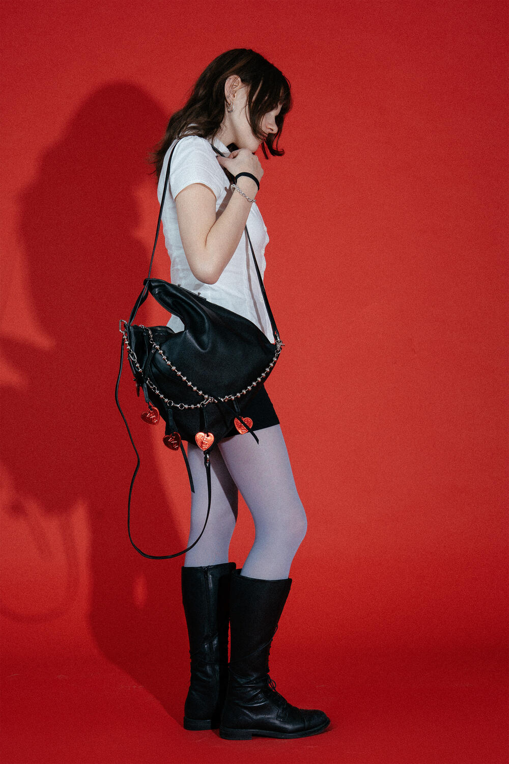 A person in a white t-shirt, black shorts, white tights, and black boots stands in profile against a red background, holding an INA KENT black bag decorated with chain and heart charms.