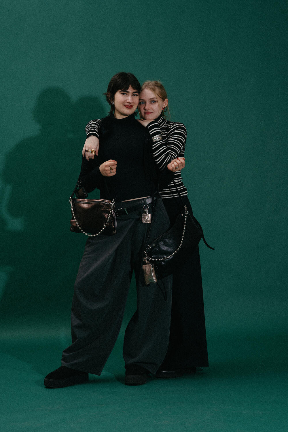 Two women stand close together against a green backdrop, both wearing dark, loose-fitting clothing and INA KENT shoulder bags with metal chains, looking at the camera.