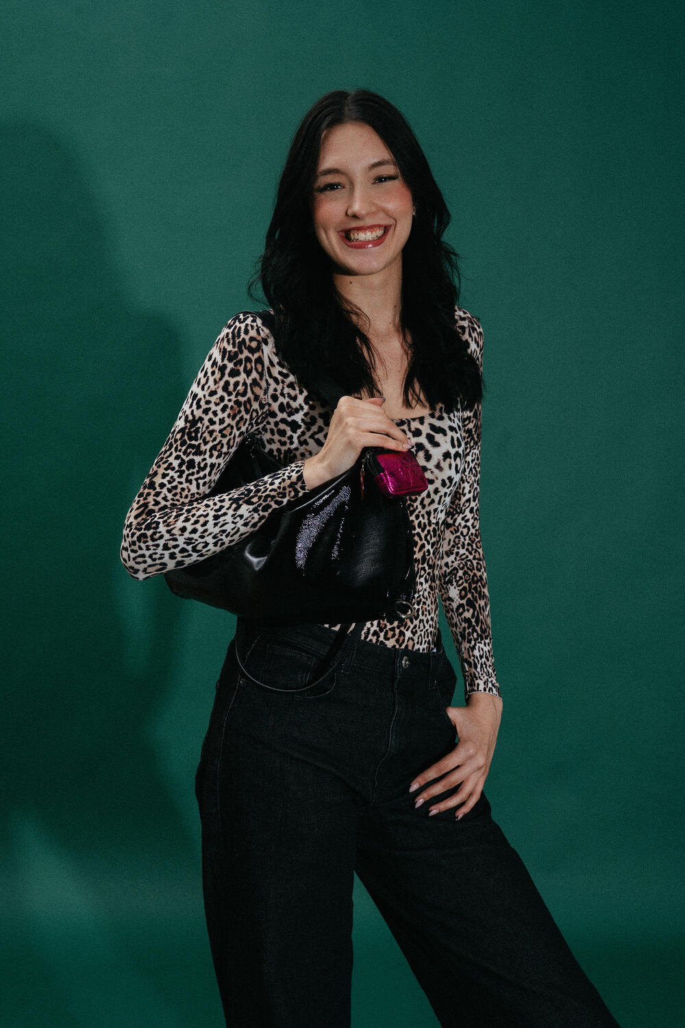 Woman wearing a leopard print top and black jeans stands against a green background, smiling and holding an INA KENT black handbag.