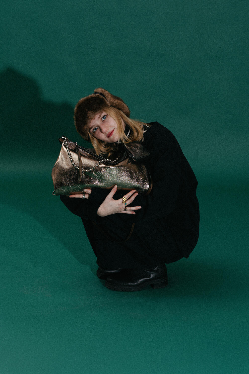 A person wearing a brown furry hat and black clothing squats on a green floor, holding a large metallic INA KENT handbag.