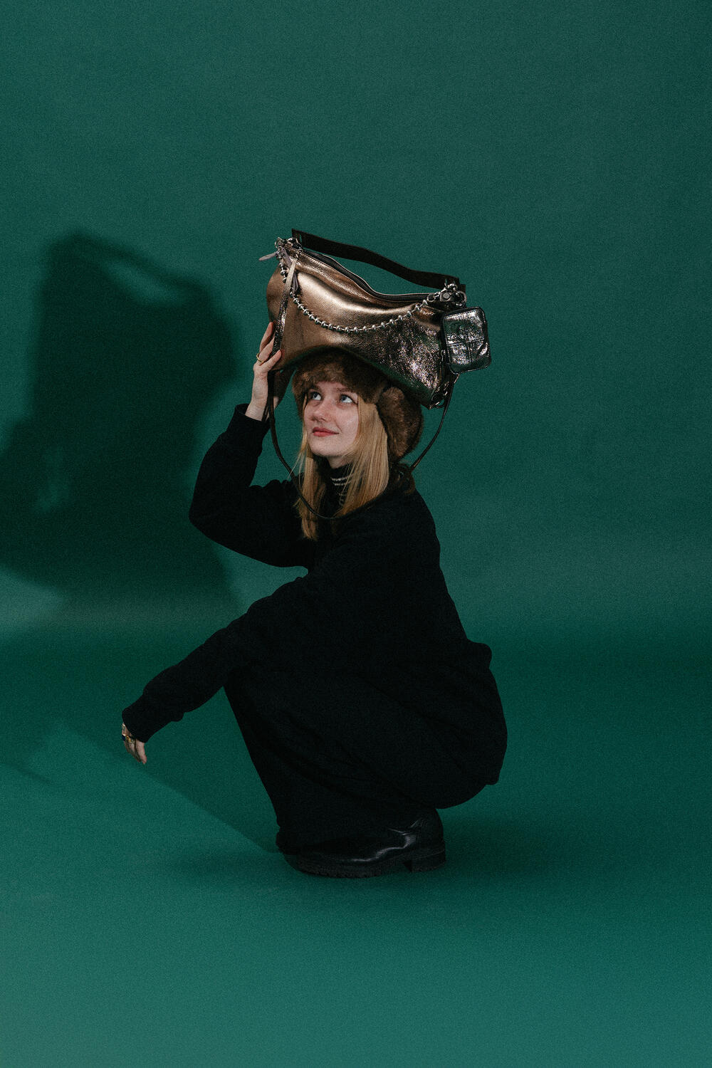 A woman in black clothing squats on the floor and balances a large metallic INA KENT handbag on her head against a dark green background.