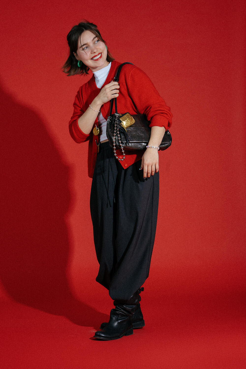 Woman standing against a red background, wearing a red sweater, white turtleneck, black wide-leg pants, black boots, and carrying an INA KENT brown shoulder bag.