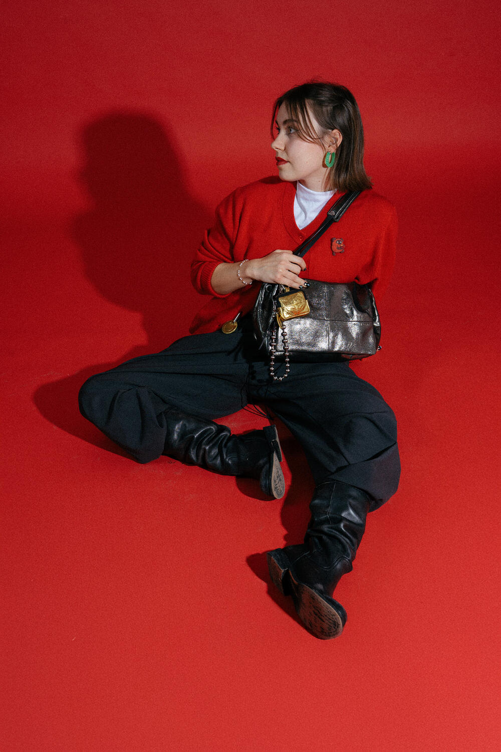 Person sitting on a red floor against a red background, wearing a red sweater, black pants, black boots, and holding an INA KENT metallic handbag.