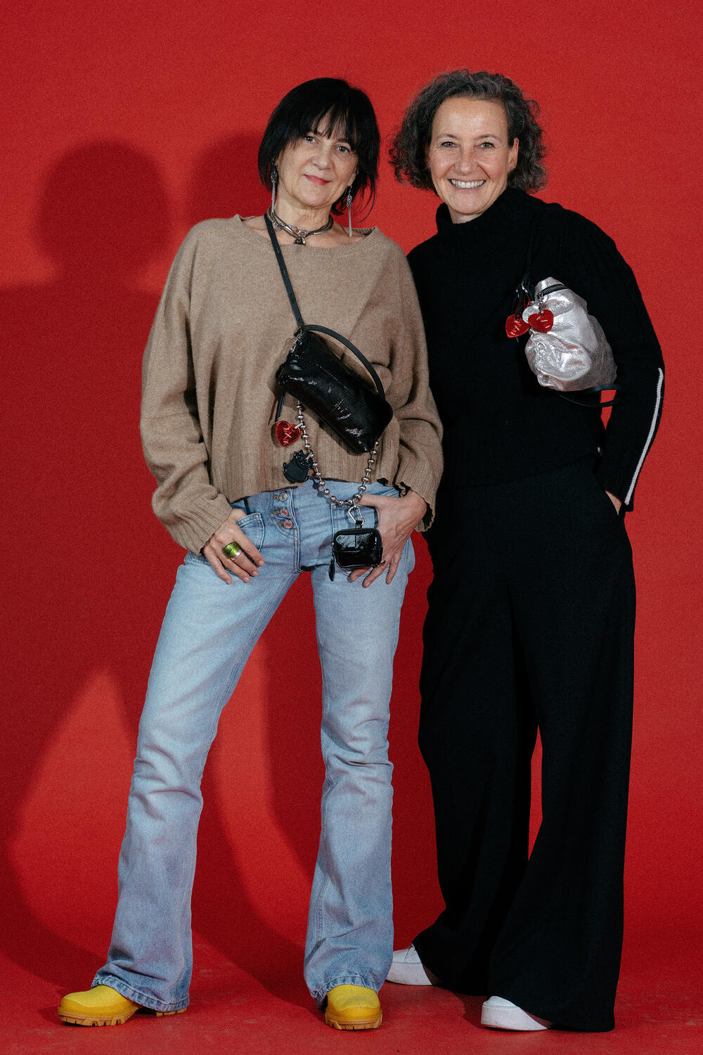 Two women stand before a red background, each carrying chic INA KENT handbags. One wears a tan sweater with blue jeans and yellow shoes; the other styles a black top and trousers.