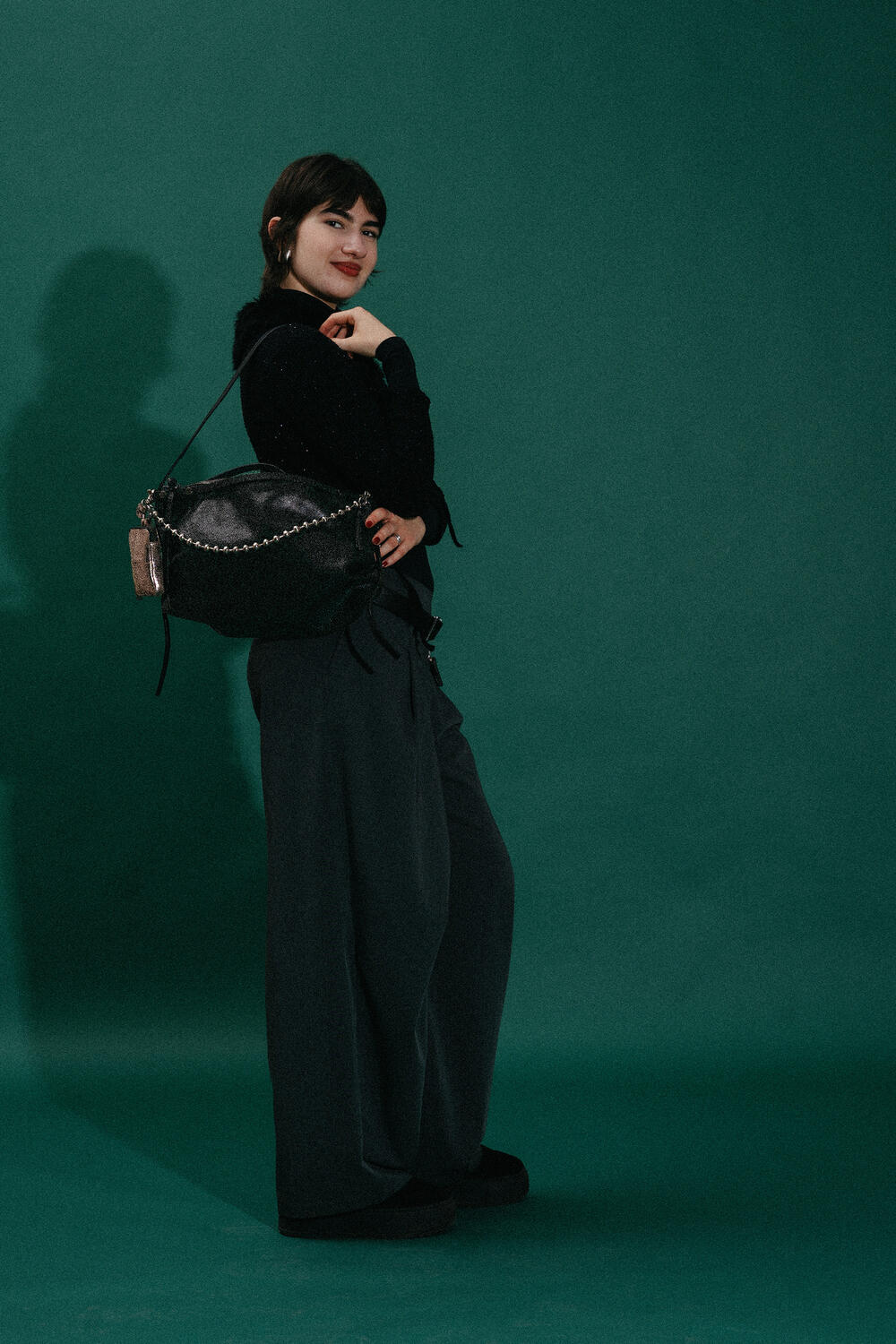A person stands in front of a green background, wearing dark wide-leg pants and a black top, carrying a large black INA KENT bag over their shoulder.