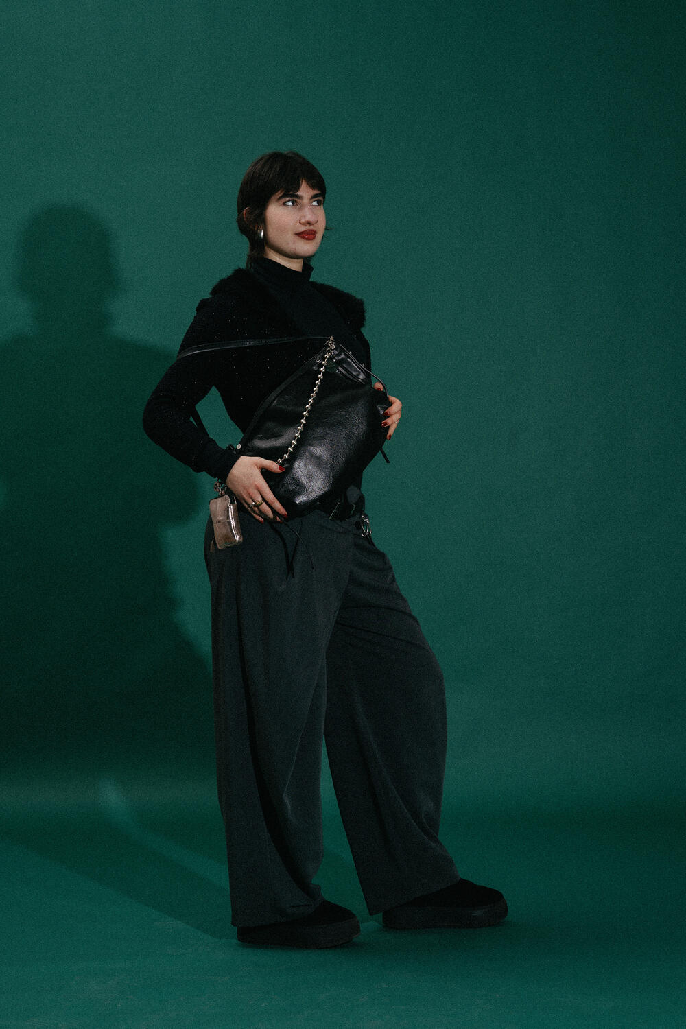 A person stands against a dark green background, wearing a black turtleneck and wide-leg pants while holding a large INA KENT black handbag.