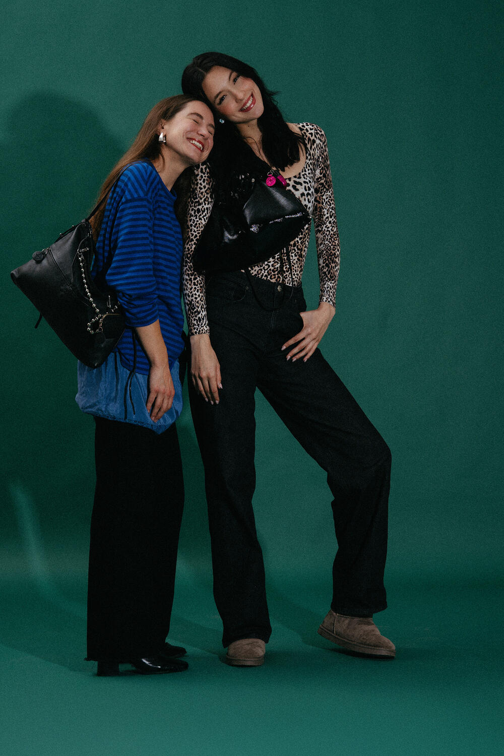 Two women stand close together against a green background, both smiling and carrying chic black INA KENT handbags. One wears a blue striped top, the other a leopard print top with black pants.