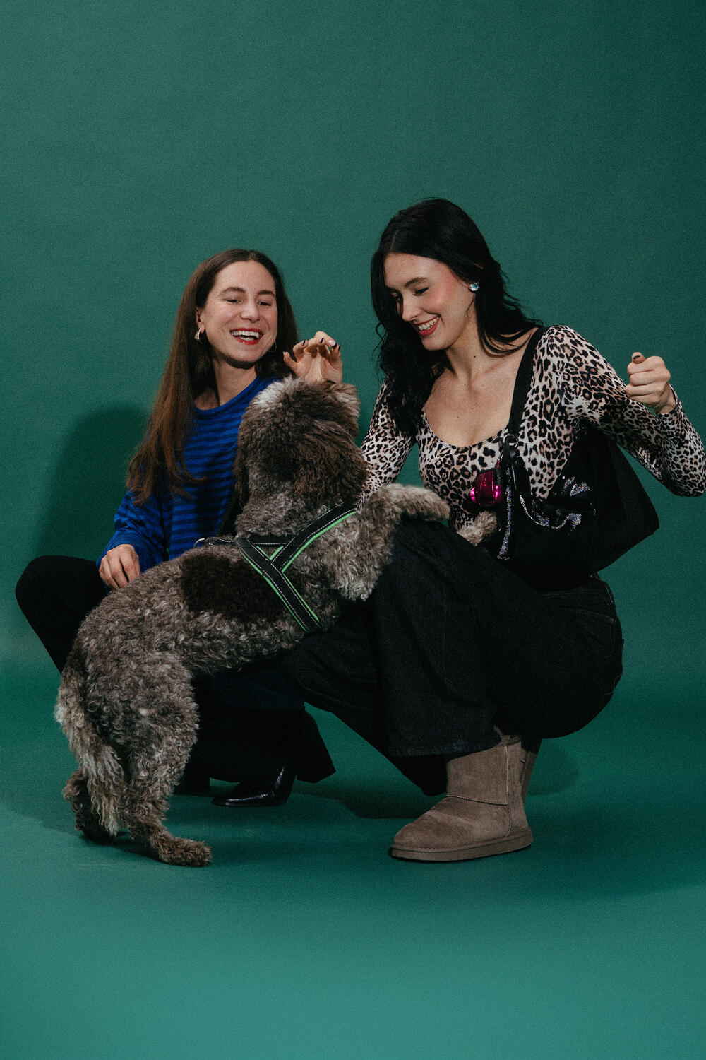 Two women smile and crouch together as a large curly-haired dog with a harness, next to an INA KENT bag, places its front paws on one of them, against a green background.