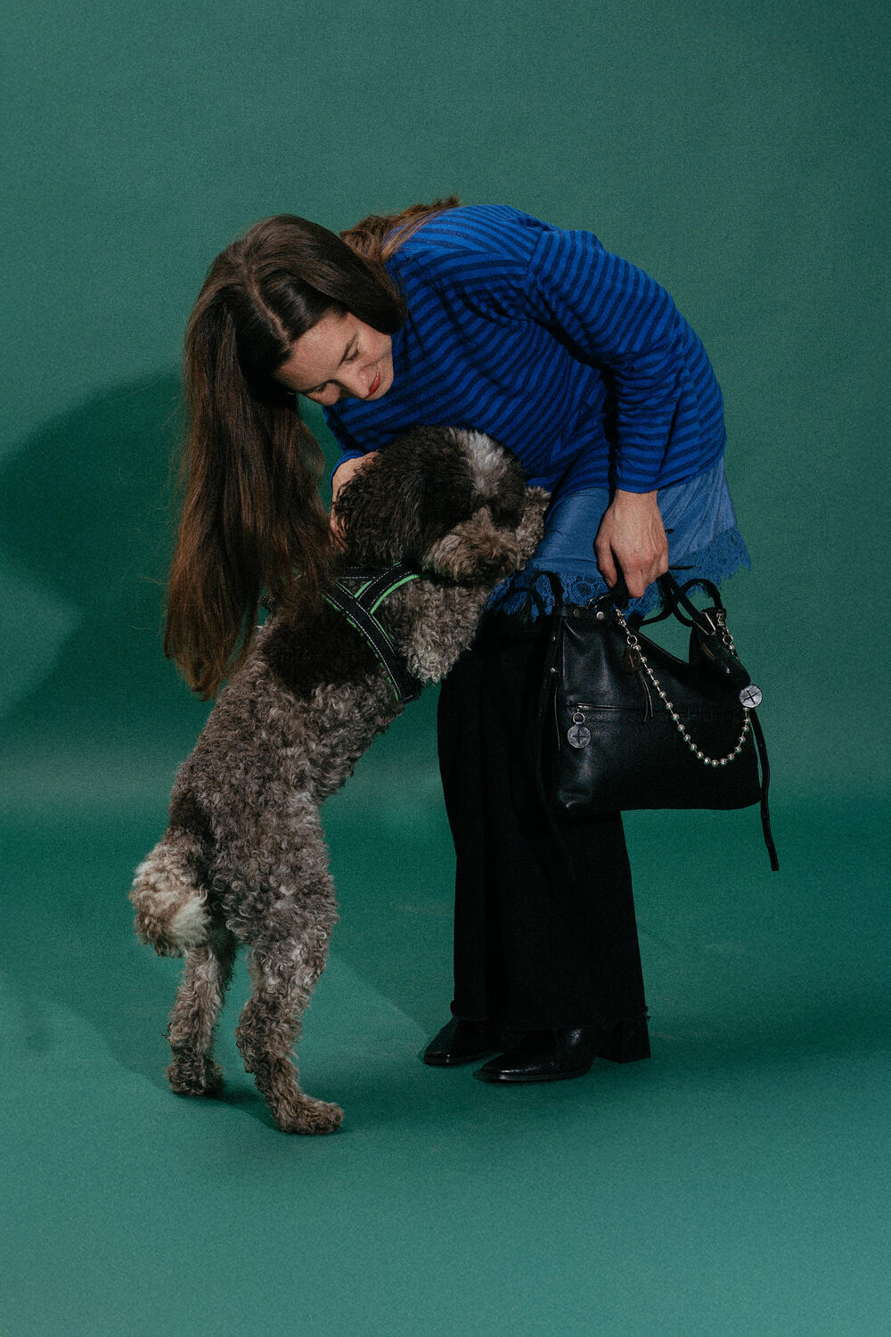 A woman in a blue striped sweater bends down to greet a large, curly-haired dog standing on its hind legs with paws on her waist. She holds an elegant black INA KENT handbag.