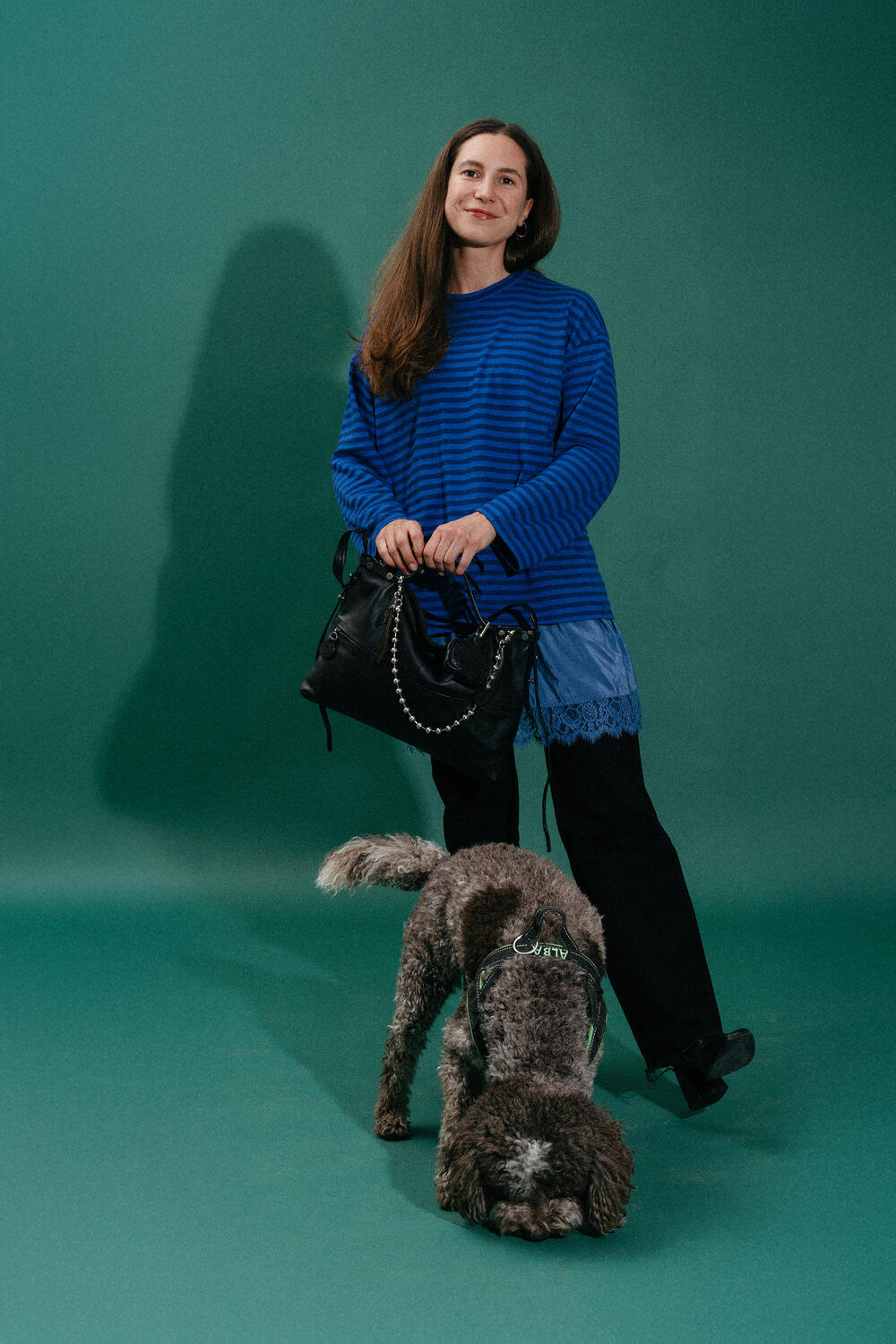 A woman in a blue striped top and black pants holds an INA KENT black handbag while standing next to a brown curly-haired service dog sniffing the floor.