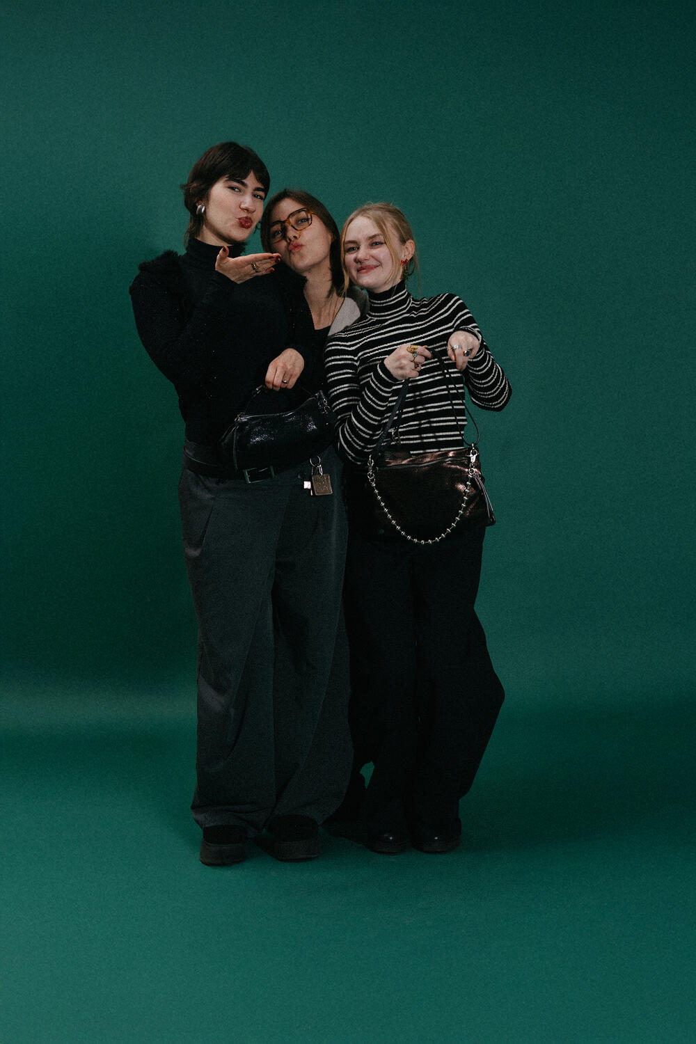 Three people stand together against a green background, posing for the camera. Two are smiling, and one is blowing a kiss. They are dressed in dark, casual INA KENT clothing.