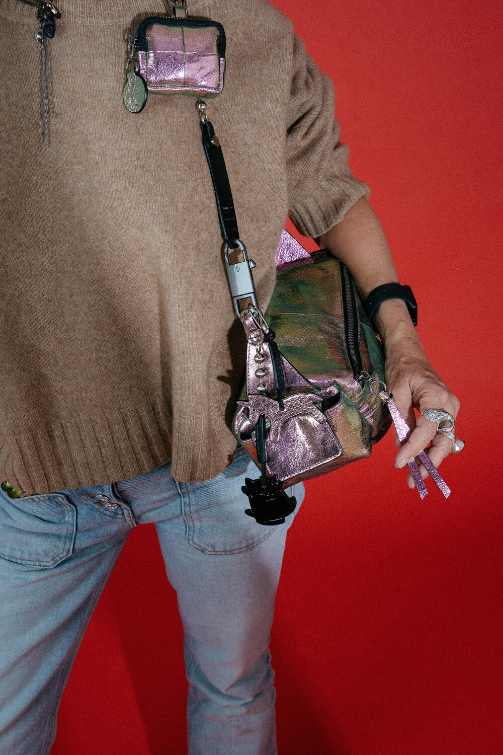 Person wearing a brown sweater and light jeans holds an INA KENT metallic iridescent bag and mini pouch against a striking red background.