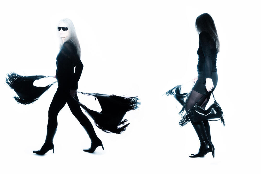 Two women in black outfits and high-heeled boots, each holding a black fringed INA KENT bag, stand and walk against a white background.