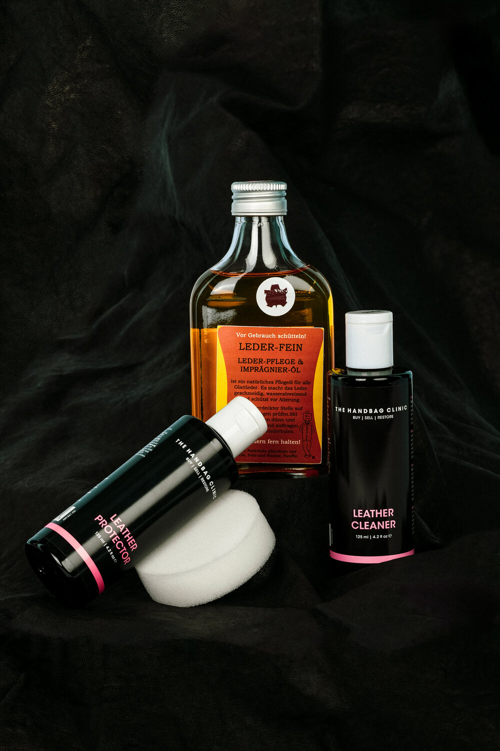 Drei INA KENT Lederpflegeprodukte - Lederöl, -reiniger und -schutz - werden mit zwei runden Schwämmen auf einem dunkel strukturierten Hintergrund dargestellt.