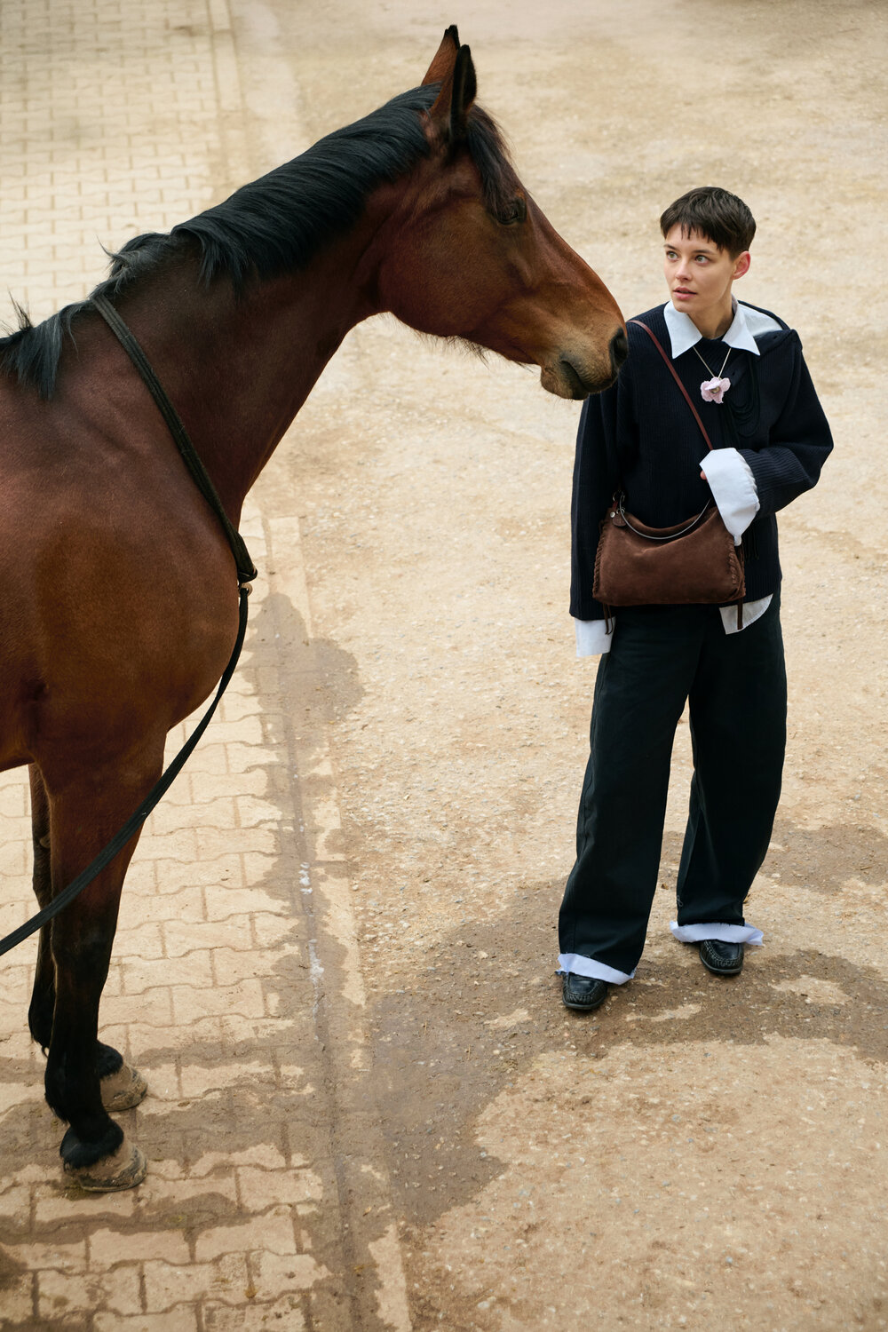 A person in loose dark clothing and an INA KENT shoulder bag stands on a paved area, facing a brown horse wearing a bridle.
