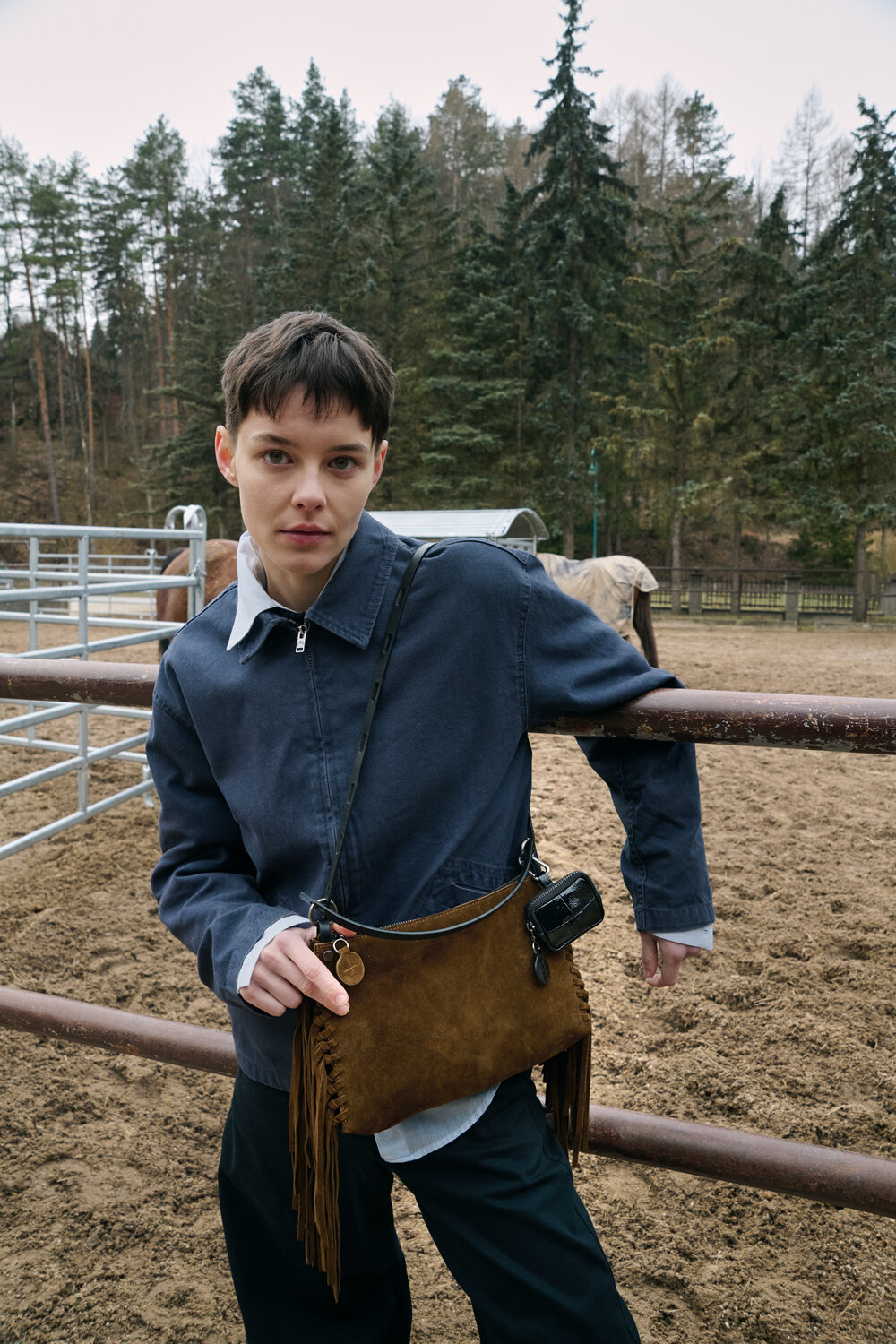 Person with short hair leans against a metal fence in an outdoor enclosure, wearing a blue jacket and holding a distinctive INA KENT brown fringed bag; trees and fencing visible in the background.