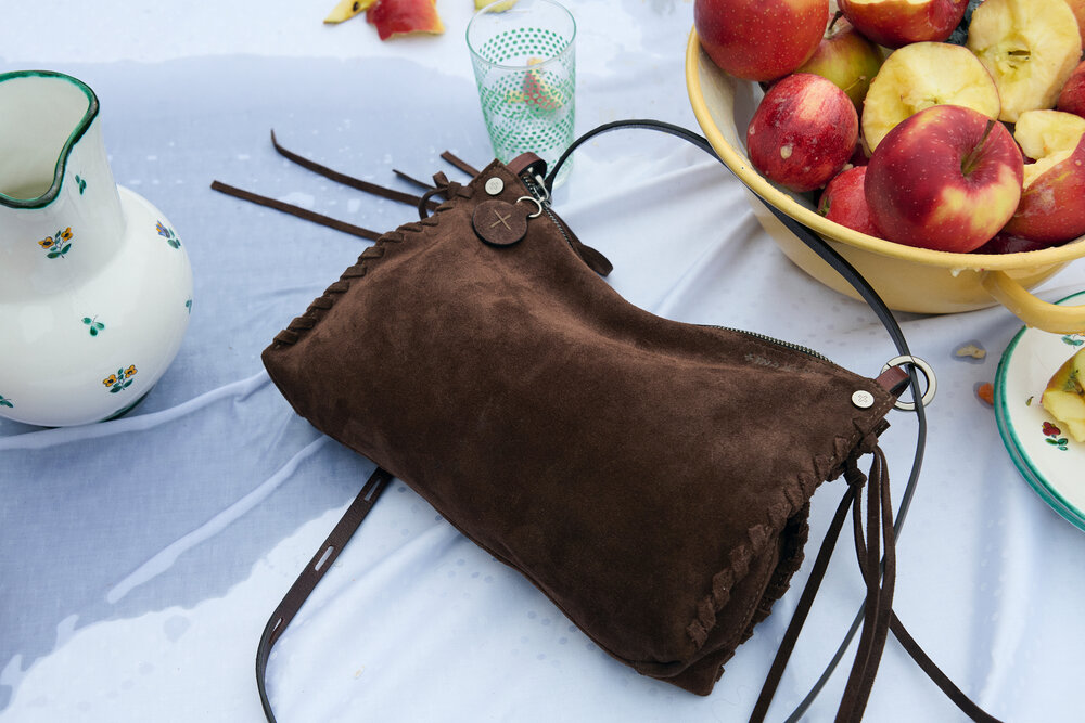 An INA KENT brown suede handbag sits on a table next to a bowl of apples, a white pitcher, and a glass with a green pattern.