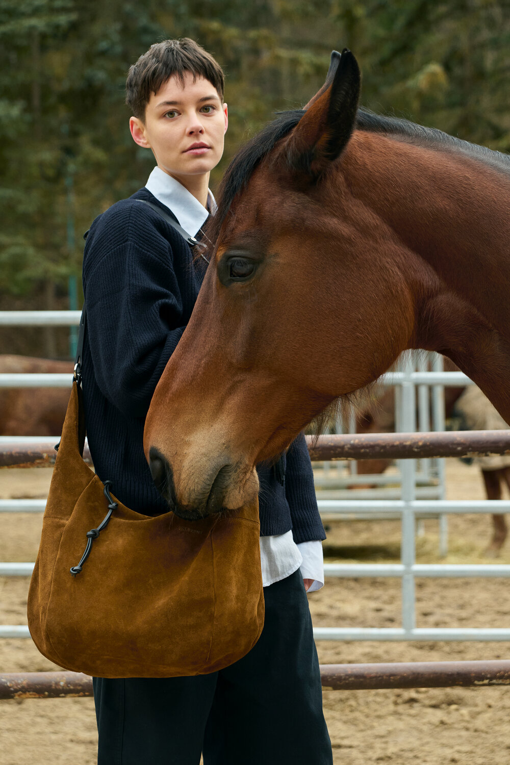 A person stands next to a brown horse that is nuzzling or sniffing an INA KENT brown suede shoulder bag in an outdoor fenced area.