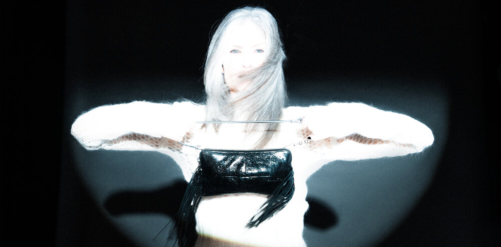 A person with long light hair holds an INA KENT black bag with fringe in front of their chest, standing in a spotlight against a dark background.