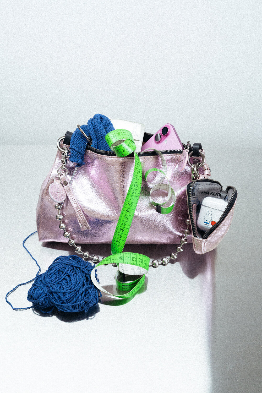 Eine rosafarbene INA KENT-Handtasche mit Strickgarn, ein blaues Strickteil, ein grünes Maßband, ein rosafarbenes Telefon und ein kleiner Beutel, ausgestellt auf einer reflektierenden Oberfläche.