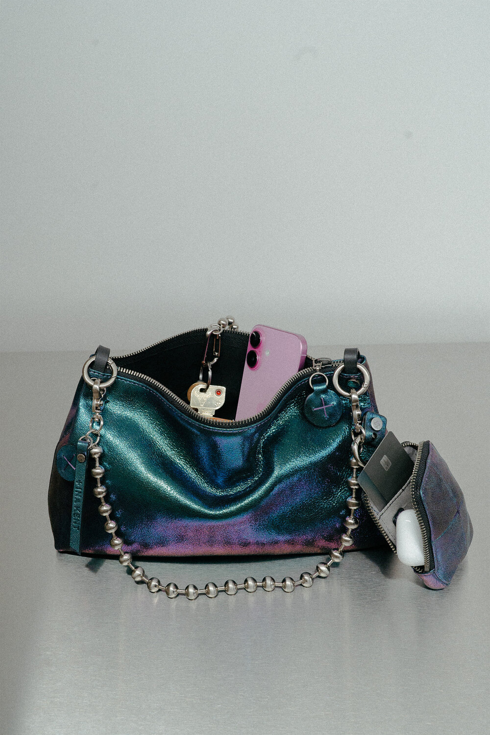 Iridescent INA KENT handbag with a metal bead strap, containing a purple smartphone, keys, and a small matching pouch holding wireless earbuds, placed on a gray surface.