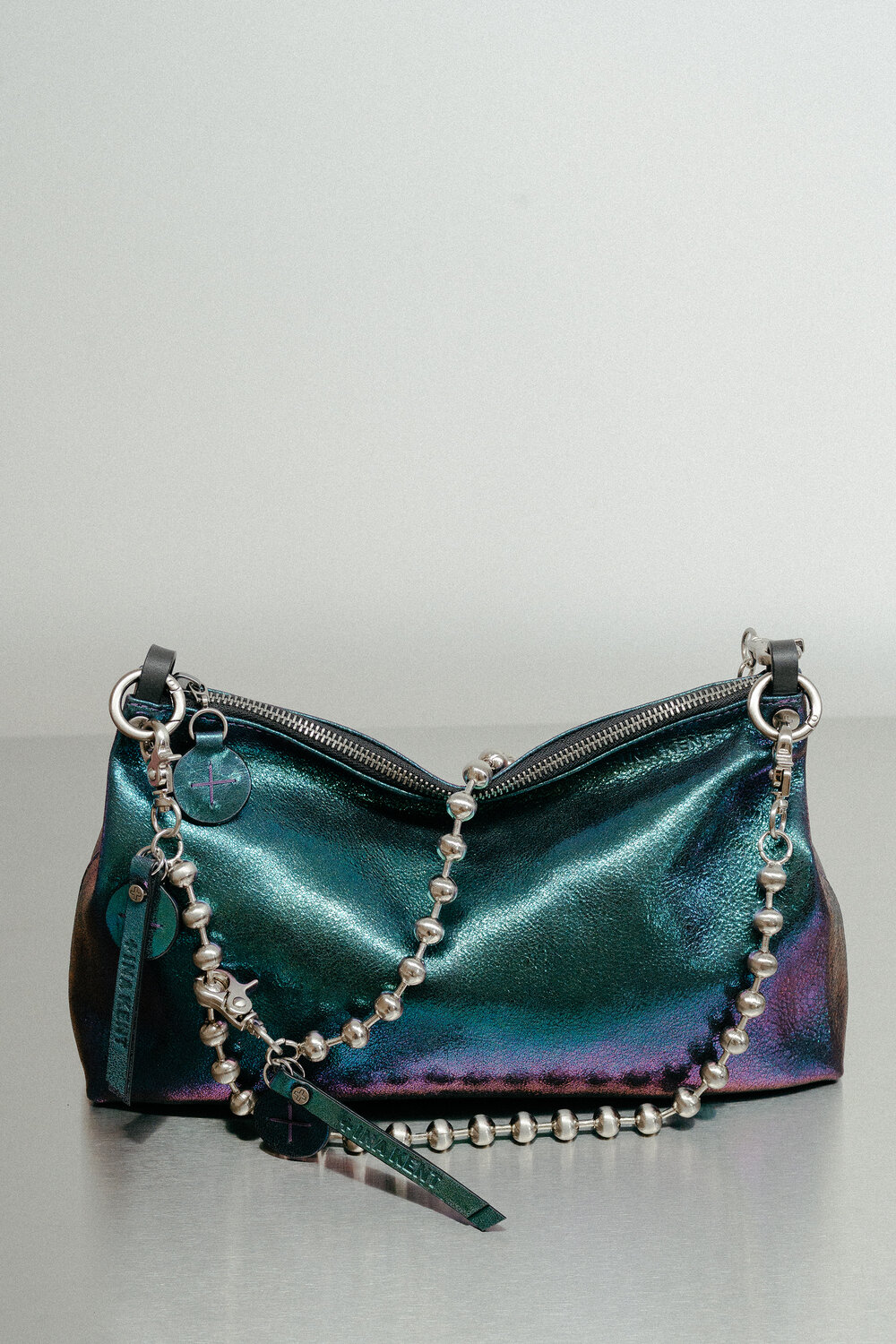A metallic, iridescent INA KENT handbag with a silver bead chain strap and zipper, displayed on a reflective surface against a plain background.