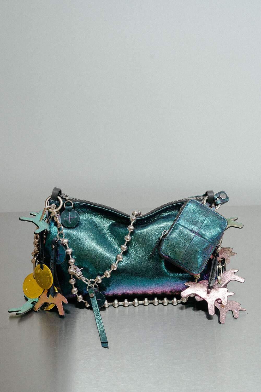 An INA KENT metallic teal handbag with a beaded strap, zipper pouch, and decorative charms—including yellow discs and abstract shapes—sits on a gray surface against a neutral background.