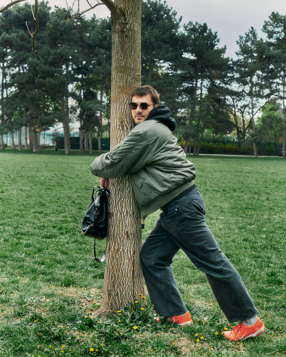 Eine Person, die eine Sonnenbrille, eine grüne Jacke, eine dunkle Hose und leuchtend orangefarbene Schuhe trägt, lehnt an einem Baum in einem grasbewachsenen Park und hält stilvoll eine schwarze INA KENT-Tasche.